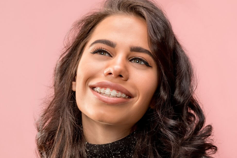 Woman with braces & wavy brown hair is smiling and looking off into the distance in front of a pink background.