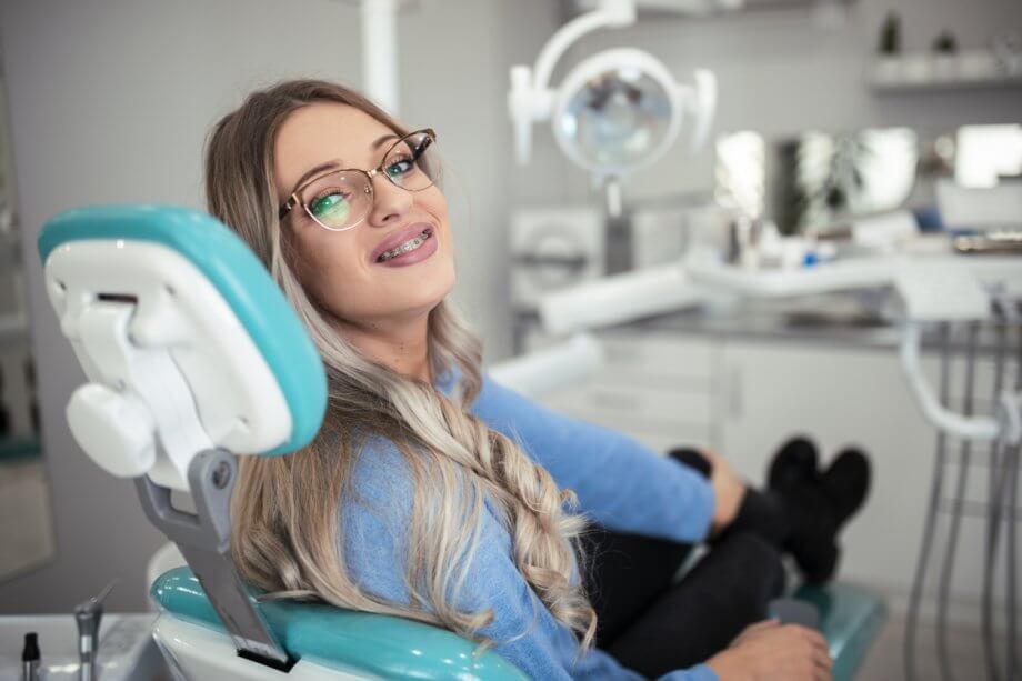 Young girl with metal braces & glasses sitting in a dentist chair and smiling.